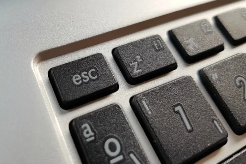 Close-up da tecla "esc" (escape) em um teclado de computador moderno, que é preta com letras brancas, contrastando com o corpo prateado do laptop. Outras teclas próximas, como "z²" e "1!", também são visíveis.