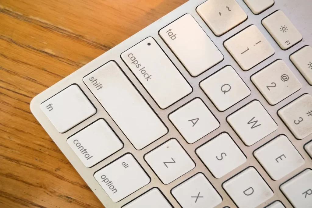 Close-up de um teclado de computador branco, focando na tecla "caps lock" e nas teclas adjacentes como "shift", "tab", "control", "option" e as letras "A", "Q", "Z", "S", "W", "X", "D", "E". A superfície da mesa de madeira é visível ao fundo.