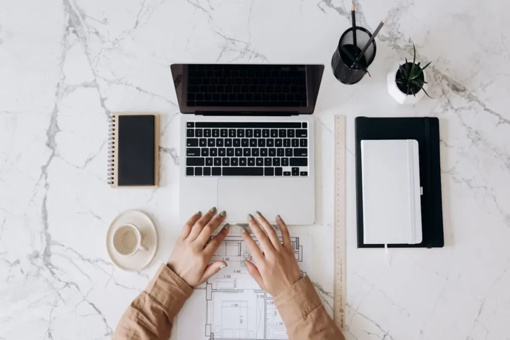 Vista superior de uma mesa com notebook, café, caderno, régua e planta decorativa, representando um ambiente de trabalho organizado