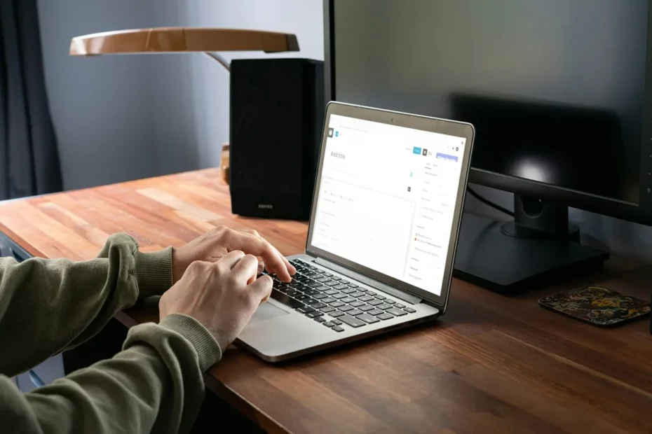 Pessoa usando moletom verde digitando em laptop sobre mesa de madeira em ambiente de home office, com monitor desligado ao fundo.