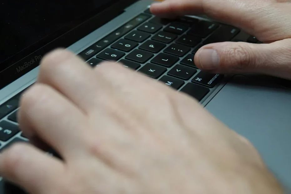 Mãos adultas digitando em teclado preto de MacBook Pro visto de perto, com foco nos dedos e teclas iluminadas.
