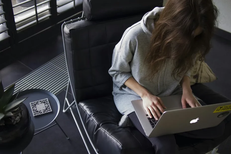 Mulher com moletom cinza digitando em notebook Apple enquanto está sentada em poltrona preta acolchoada, em ambiente interno moderno.