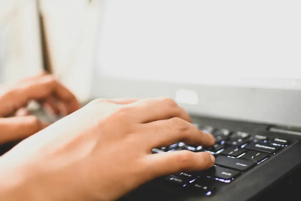 Mãos de uma pessoa digitando em um teclado preto de notebook com tela clara ao fundo, em ambiente iluminado.