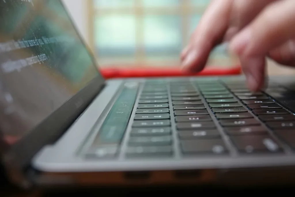 Dedos digitando em teclado de notebook visto de perto, com tela parcialmente visível e ambiente desfocado ao fundo.