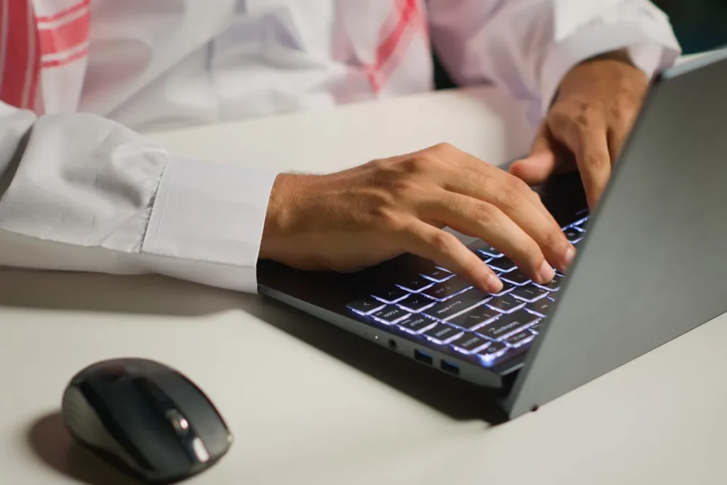 "Mão digitando em laptop com teclado iluminado e mouse ao lado, em ambiente noturno