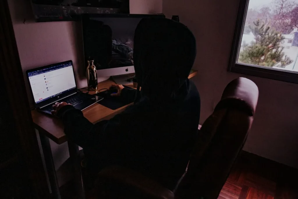 Pessoa encapuzada usando computador em ambiente escuro, com múltiplas telas e janela ao fundo.