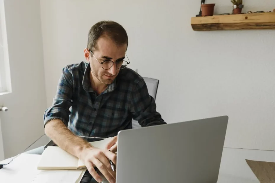 Um homem branco, de óculos redondos e camisa xadrez escura, sentado em uma mesa branca, concentrado em seu laptop prateado. Ele tem um caderno aberto e uma caneta próximos, e parece estar anotando algo ou estudando. Ao fundo, uma parede branca e uma prateleira de madeira com pequenos vasos.