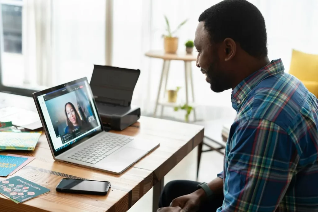 Homem sorridente usando camisa xadrez participa de entrevista online por videoconferência em um notebook, com uma mulher aparecendo na tela