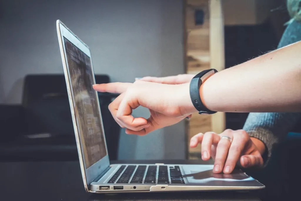 Duas mãos interagindo com notebook, uma apontando para a tela e outra digitando no teclado