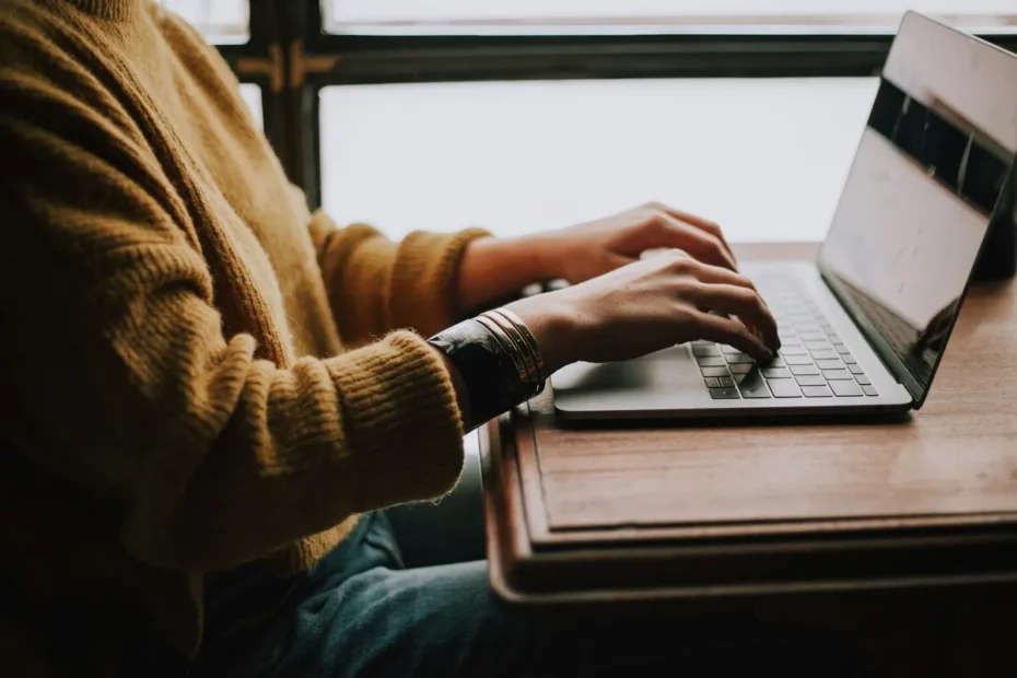 Pessoa com suéter amarelo digitando em notebook em uma mesa de madeira perto de uma janela