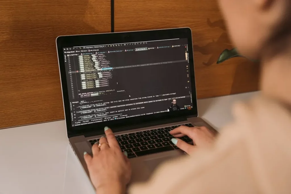 Visão por cima do ombro de uma pessoa digitando em um laptop com código de programação visível na tela. As mãos da pessoa, com unhas pintadas de azul claro, estão sobre o teclado. O código na tela é escuro com texto colorido, indicando linhas de código e possíveis mensagens de erro.