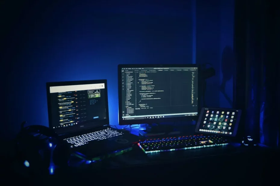 Estação de trabalho com iluminação azul, exibindo três telas: laptop Dell, monitor com código em Visual Studio Code e tablet com interface de aplicativos.