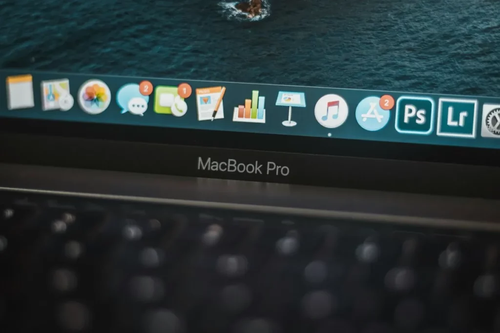 Close-up da tela de um MacBook Pro com dock do macOS exibindo diversos aplicativos, incluindo Photoshop e App Store.