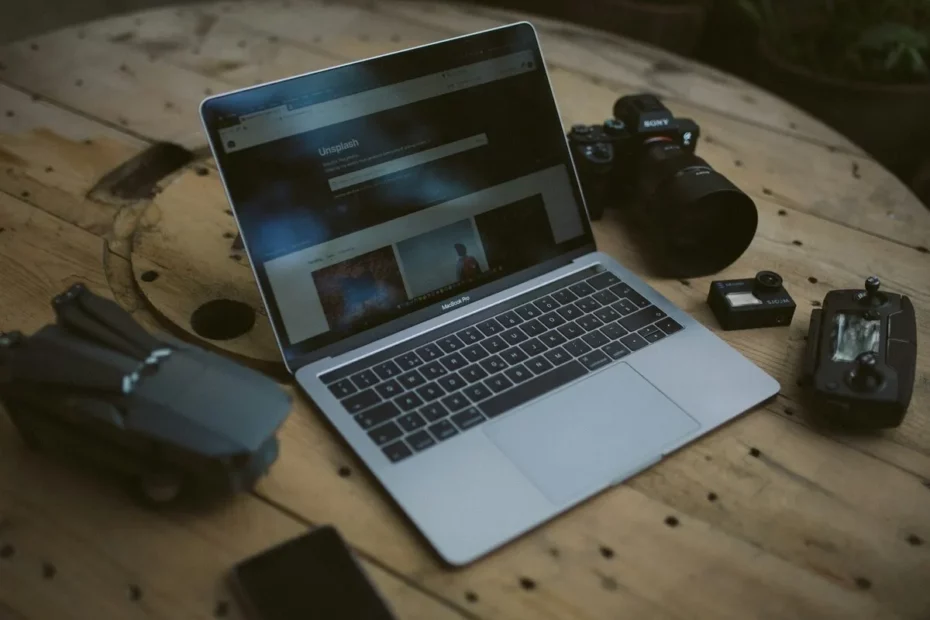 Notebook MacBook Pro com site da Unsplash aberto, ao lado de câmera Sony, drone e acessórios sobre uma mesa de madeira