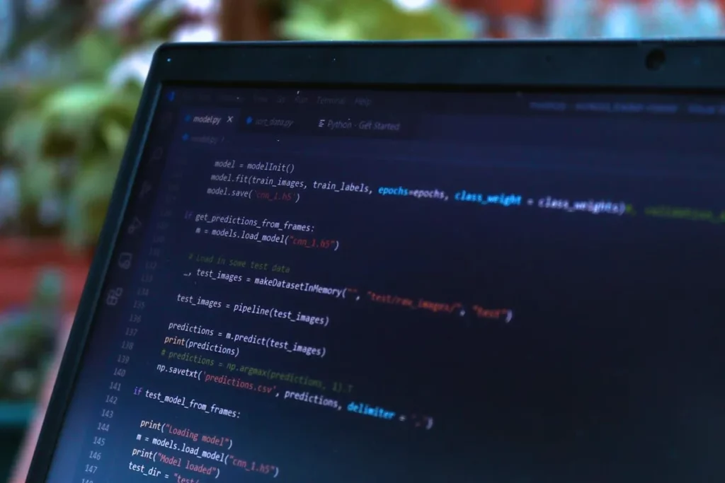Profissões em alta: Close-up de uma tela de laptop exibindo linhas de código de programação em um editor de texto escuro. O código parece ser em Python, com termos como "model.fit", "predictions", e "tensorflow". A interface mostra abas de arquivos e um terminal.