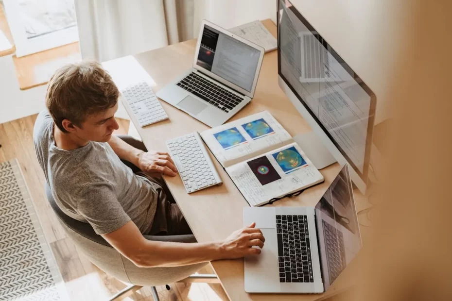 Profissões em alta: Visão superior de um jovem adulto, de cabelos claros e camiseta cinza, sentado em uma cadeira em uma mesa de madeira clara, trabalhando com um setup de múltiplos monitores. Há um laptop aberto e um monitor externo exibindo código ou dados, outro laptop aberto mais próximo do espectador, dois teclados sem fio e um caderno aberto com gráficos ou diagramas.