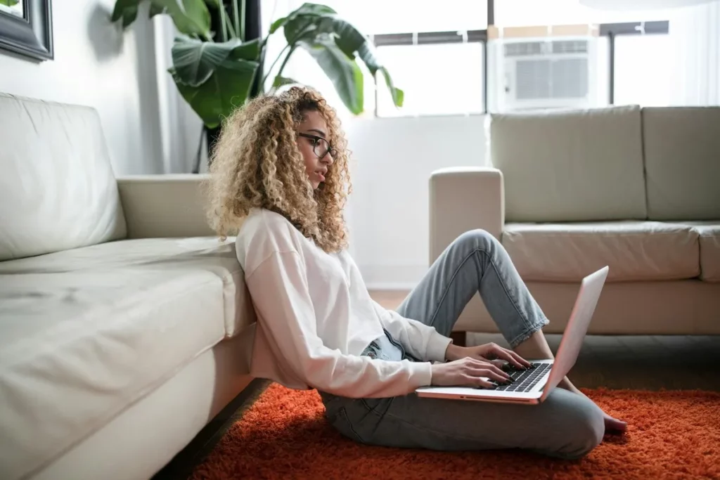 Jovem mulher com cabelos cacheados e óculos usando um laptop no chão de uma sala de estar, provavelmente buscando como liberar espaço no PC.