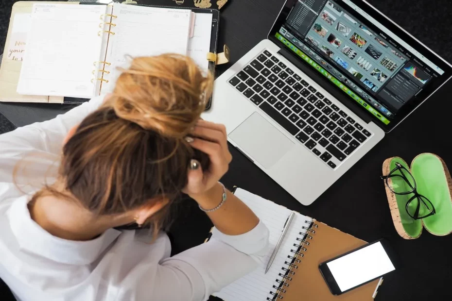 Pessoa frustrada com as mãos na cabeça em frente a um laptop e materiais de trabalho, simbolizando a necessidade de recuperar arquivos apagados.