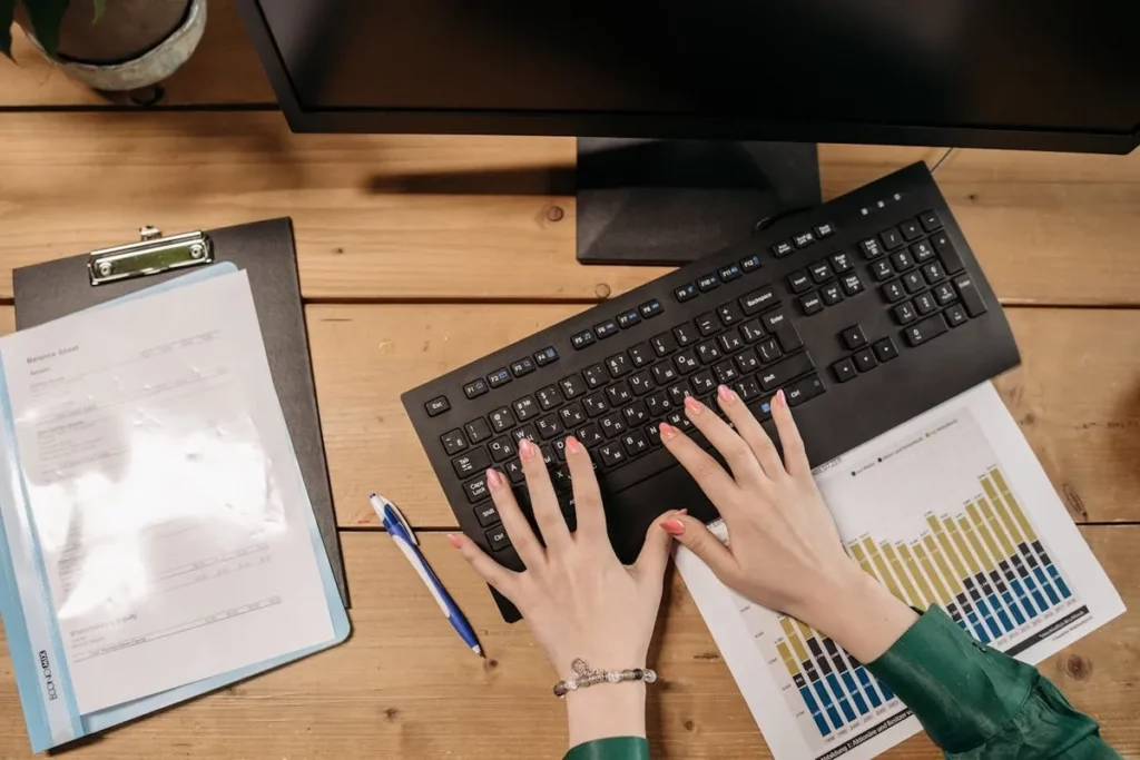 Vista superior de uma pessoa digitando em um teclado preto em uma mesa de madeira, com um monitor, uma prancheta com documentos e um gráfico de barras impresso ao lado.