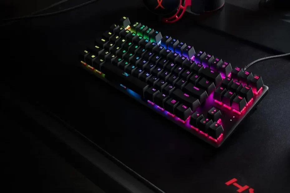 Teclado RGB mecânico preto com retroiluminação multicolorida intensa em setup gamer, sobre mousepad escuro com fone de ouvido ao fundo.