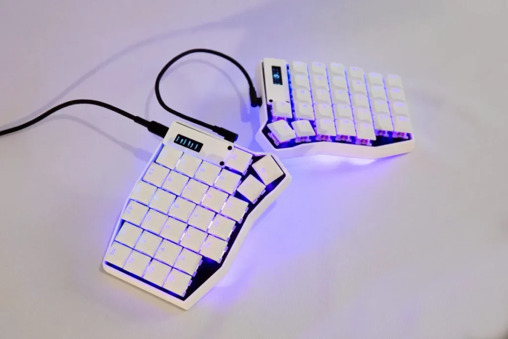 Imagem de um par de teclados split na cor branca, com iluminação de fundo LED azul e roxa. As duas metades estão separadas e conectadas por cabos pretos.