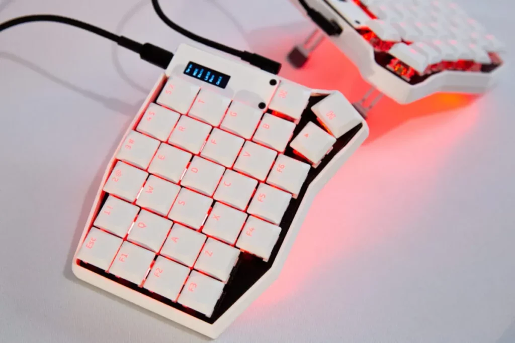 Metade esquerda de um teclado split mecânico na cor branca, com iluminação de fundo vermelha e uma pequena tela na parte superior, conectado por um cabo preto.