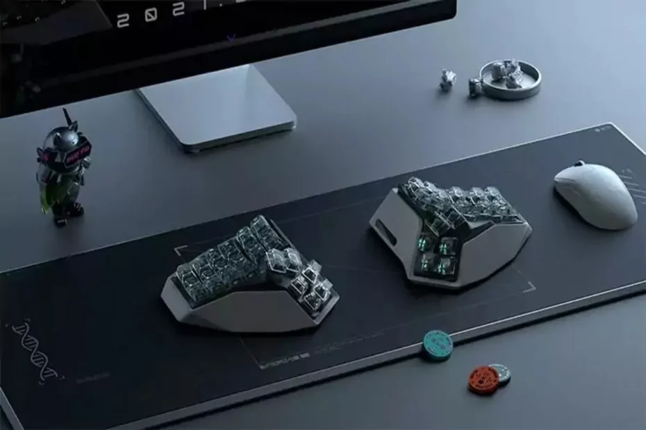 Visão de cima de um setup de trabalho minimalista com um par de teclados split brancos e transparentes, um mouse sem fio branco e um monitor em um ambiente de mesa cinza.