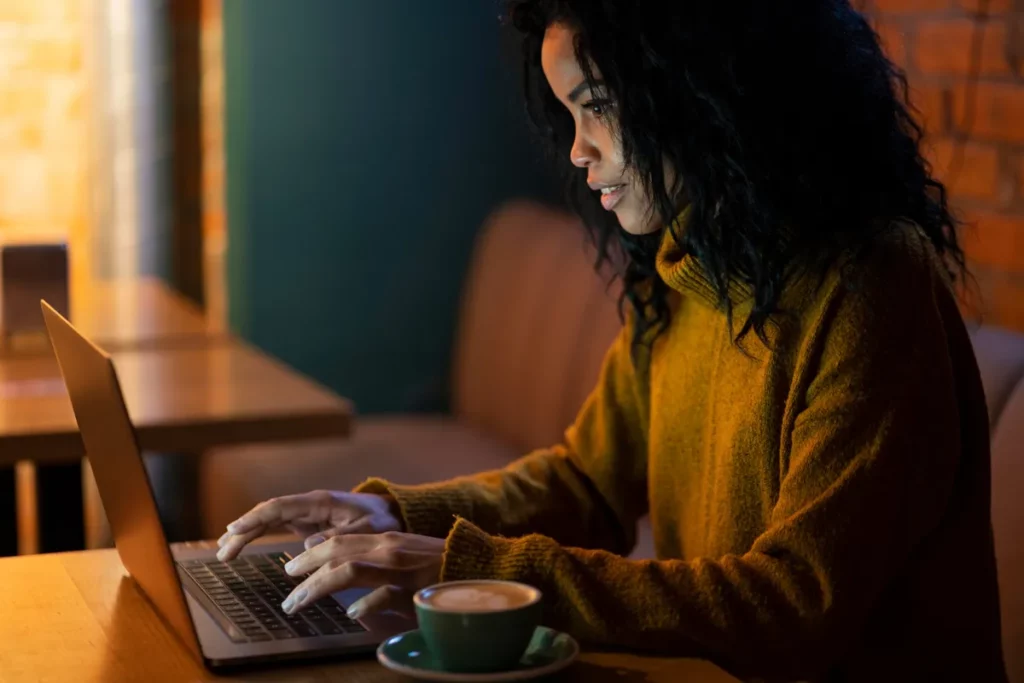 Mulher afrodescendente concentrada, digitando em seu laptop em um café ou escritório aconchegante, sugerindo foco em um jogo de digitação ou trabalho noturno.