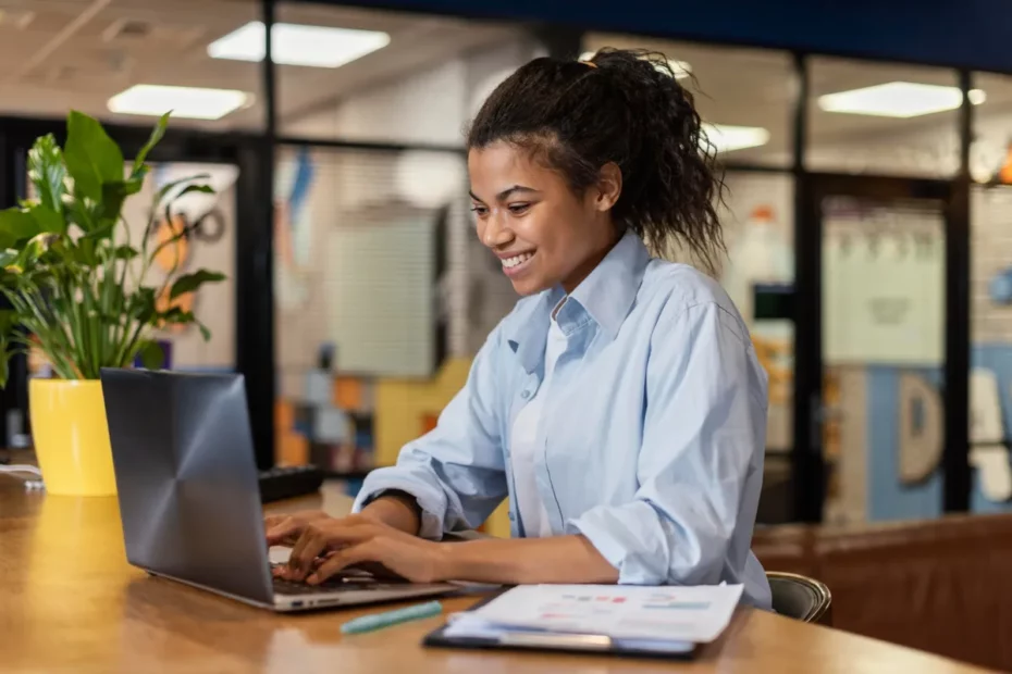 Jovem mulher sorridente, com cabelo preso, digitando em seu laptop em uma mesa de trabalho ou em um jogo de digitação, em um escritório moderno.