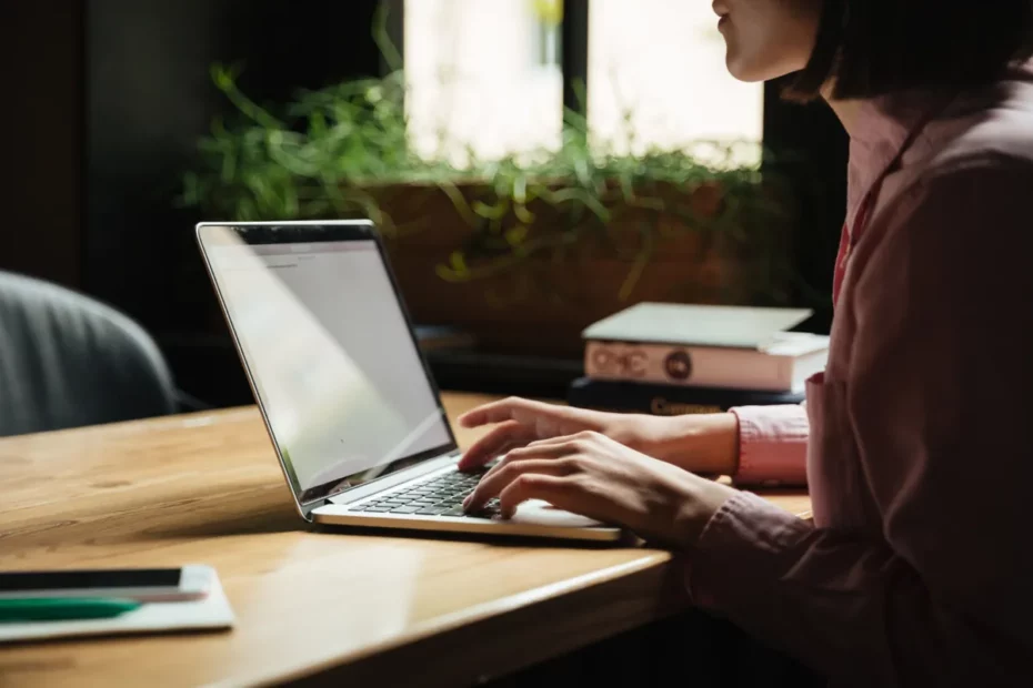 Mulher digitando em um laptop prateado sobre uma mesa de madeira, focando no trabalho de escrita e edição, com um vaso de plantas e livros no fundo, representando o uso de ferramentas como o LanguageTool.