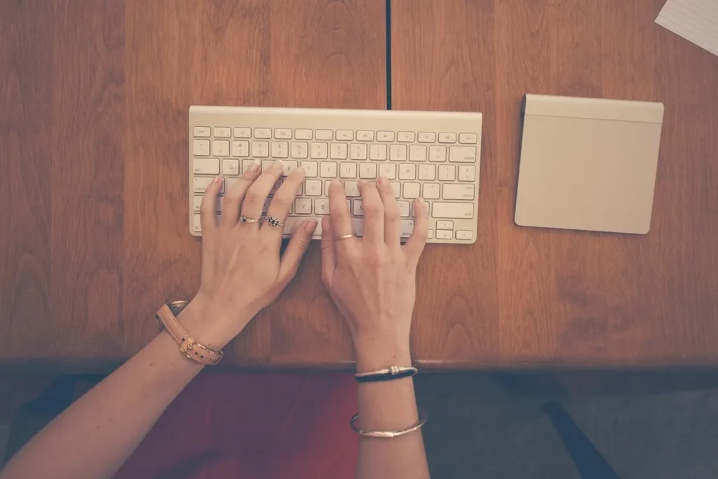 Uma visão de cima (flat lay) das mãos de uma pessoa digitando em um teclado sem fio branco, com um trackpad ao lado, sobre uma mesa de madeira. A pessoa usa anéis, pulseiras e um relógio. O foco nas mãos em ação e na organização do espaço de trabalho sugere a busca por eficiência e pequenos ajustes de rotina, pilares do Método Kaizen para otimização de processos.