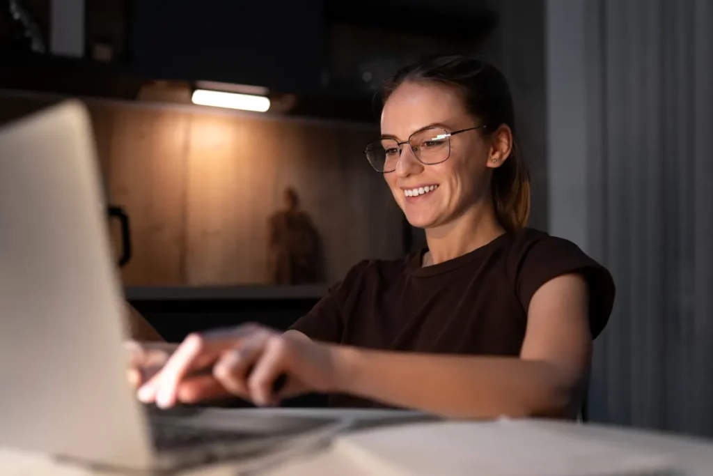 Close-up de mulher sorridente com óculos, digitando ativamente em um laptop, praticando um teste de digitação sem acentos ou trabalhando à noite.