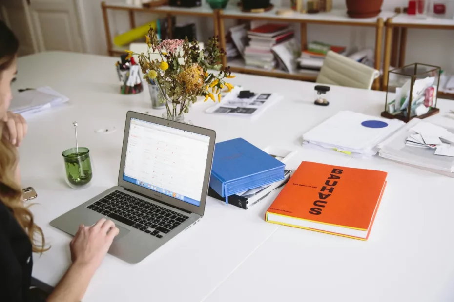 Pessoa usando um notebook em uma mesa de escritório com livros, pastas, papéis e um arranjo de flores ao fundo.