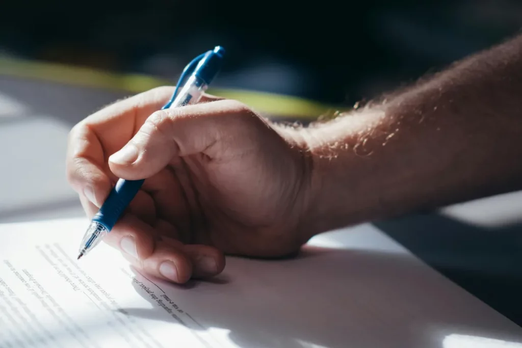 Close de uma mão segurando uma caneta azul enquanto escreve em um documento impresso.