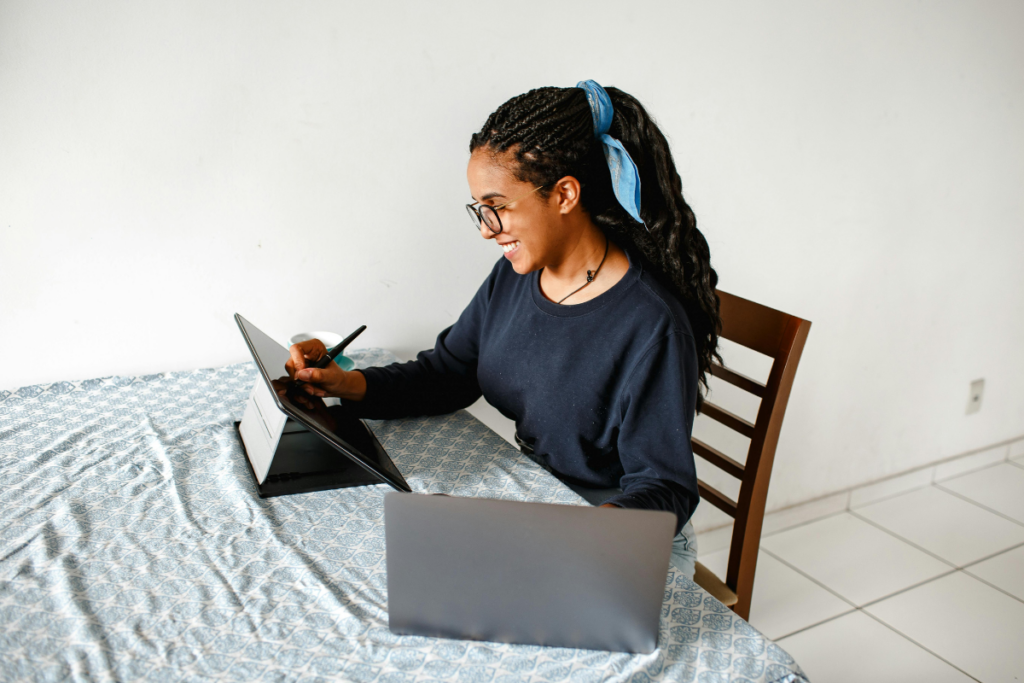Mulher sorrindo enquanto escreve com caneta digital em um tablet apoiado sobre a mesa, com um notebook aberto ao lado.
