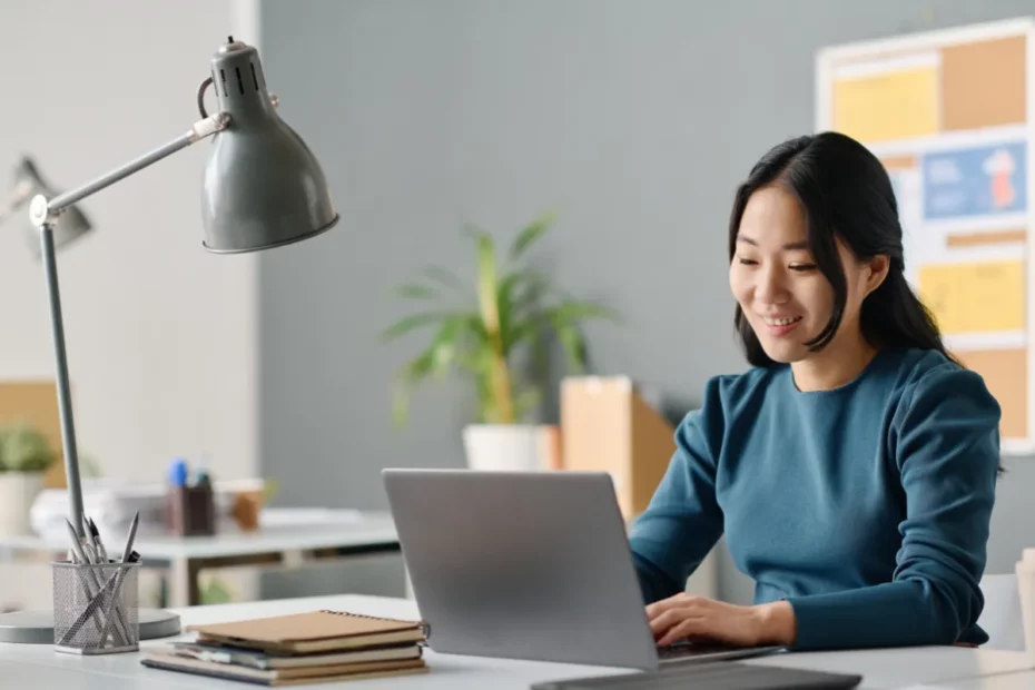 Mulher sorrindo enquanto trabalha no notebook em mesa organizada com luminária e planta ao lado.