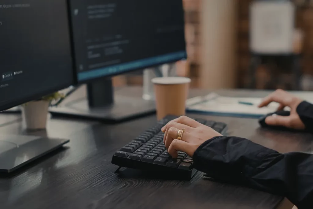 Pessoa digitando em teclado preto conectado a computador com dois monitores em mesa de trabalho